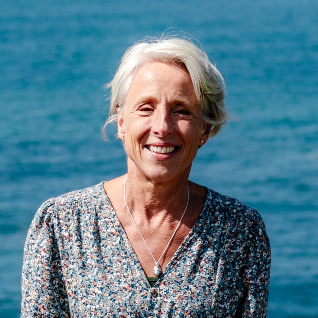 Mary stands smiling, bathed in sun, in front of the ocean.